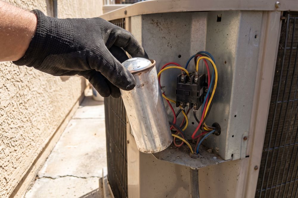 Gloved hand lifting a swollen run capacitor out of an outdoor AC unit's electrical panel