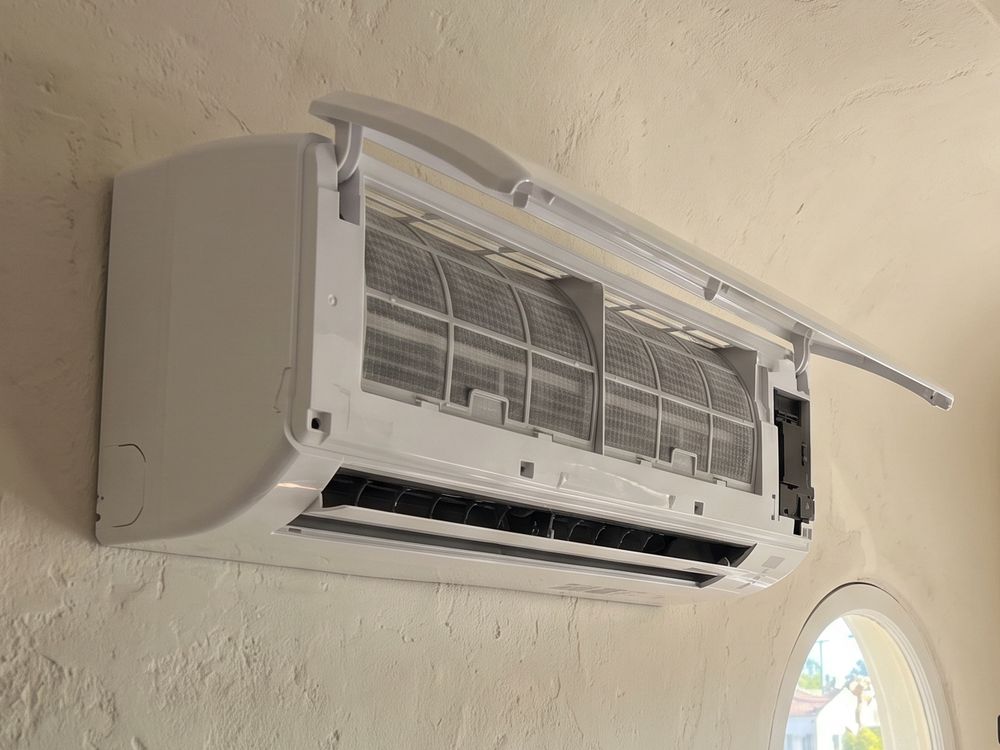 Mini-split head with its front cover lifted and filters showing, on a plaster wall beside an arched window
