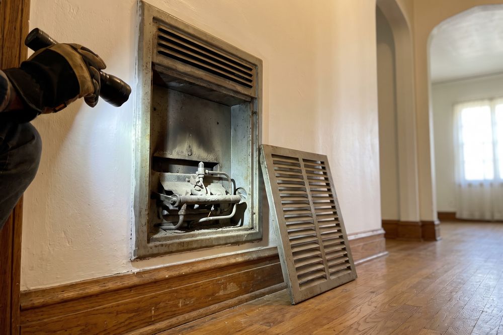 Gas wall furnace in a 1940s hallway with its grille off and a flashlight on the burner