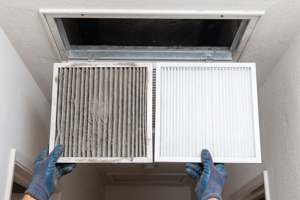 Gloved hands holding a dust-clogged air filter beside a clean one under a ceiling return grille